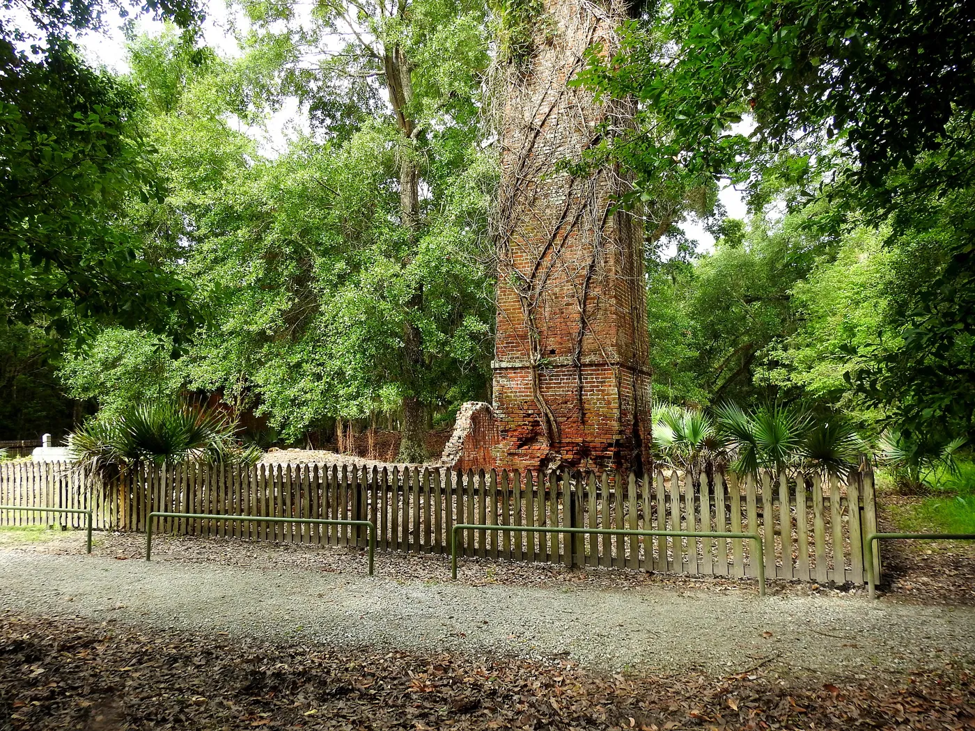 A tall crumbling brick chimney among green trees and palmettos, behind a low wooden fence