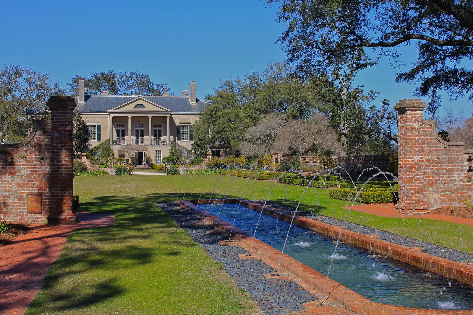 A long rectangular pool with a row of small fountains leading across a lawn to a columned white house, between brick pillars