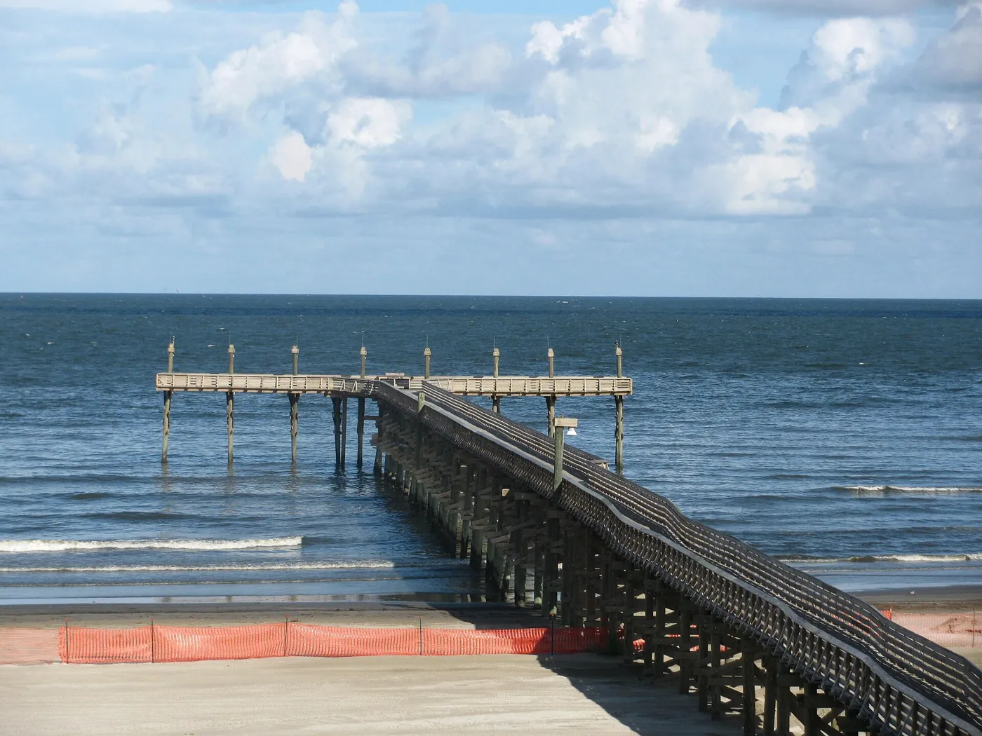 A long wooden fishing pier running out from a sandy beach into the grey-blue Gulf under a sky full of white clouds