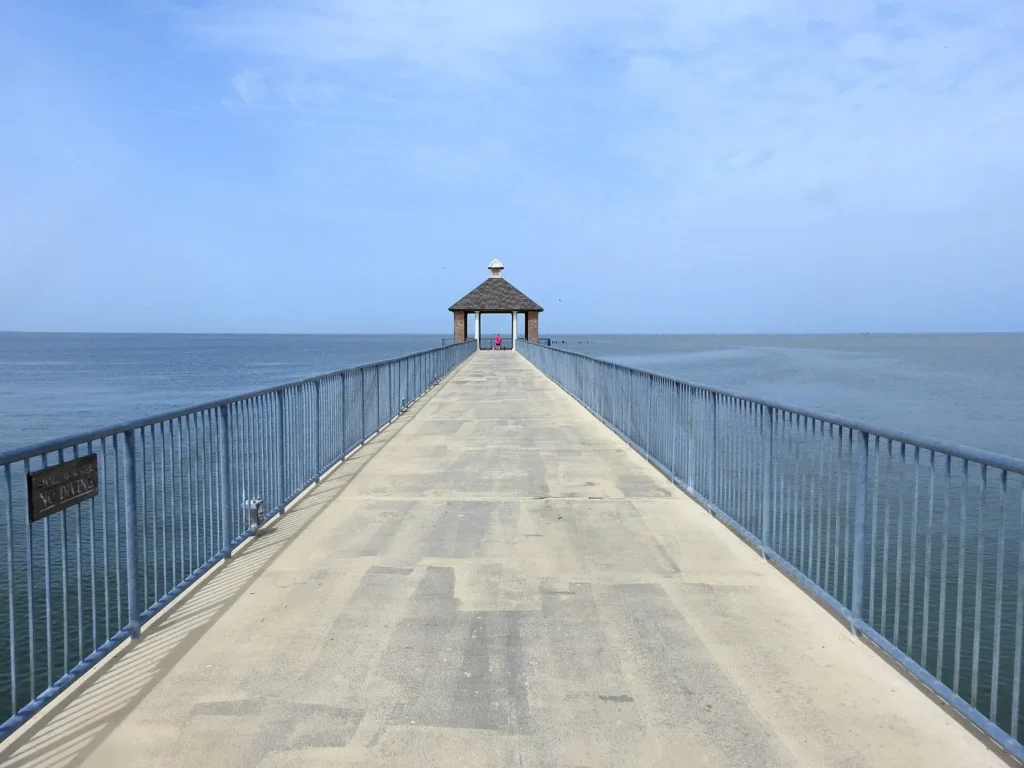 A long concrete pier with railings running straight out over calm blue Lake Pontchartrain to a small roofed gazebo